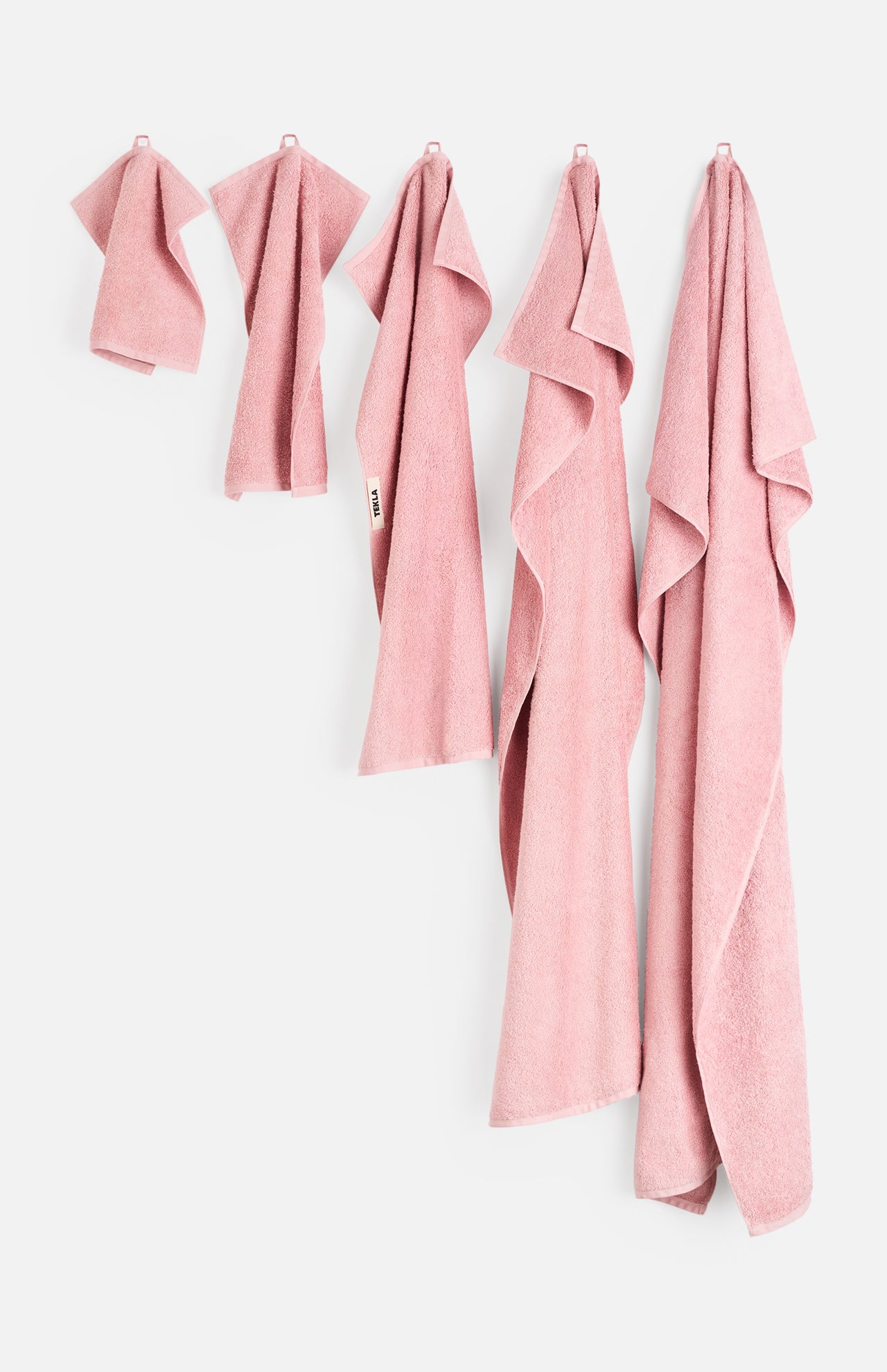 Five dusty pink organic cotton Terry Hand Towels of varying sizes hanging in a row against a white bathroom wall.