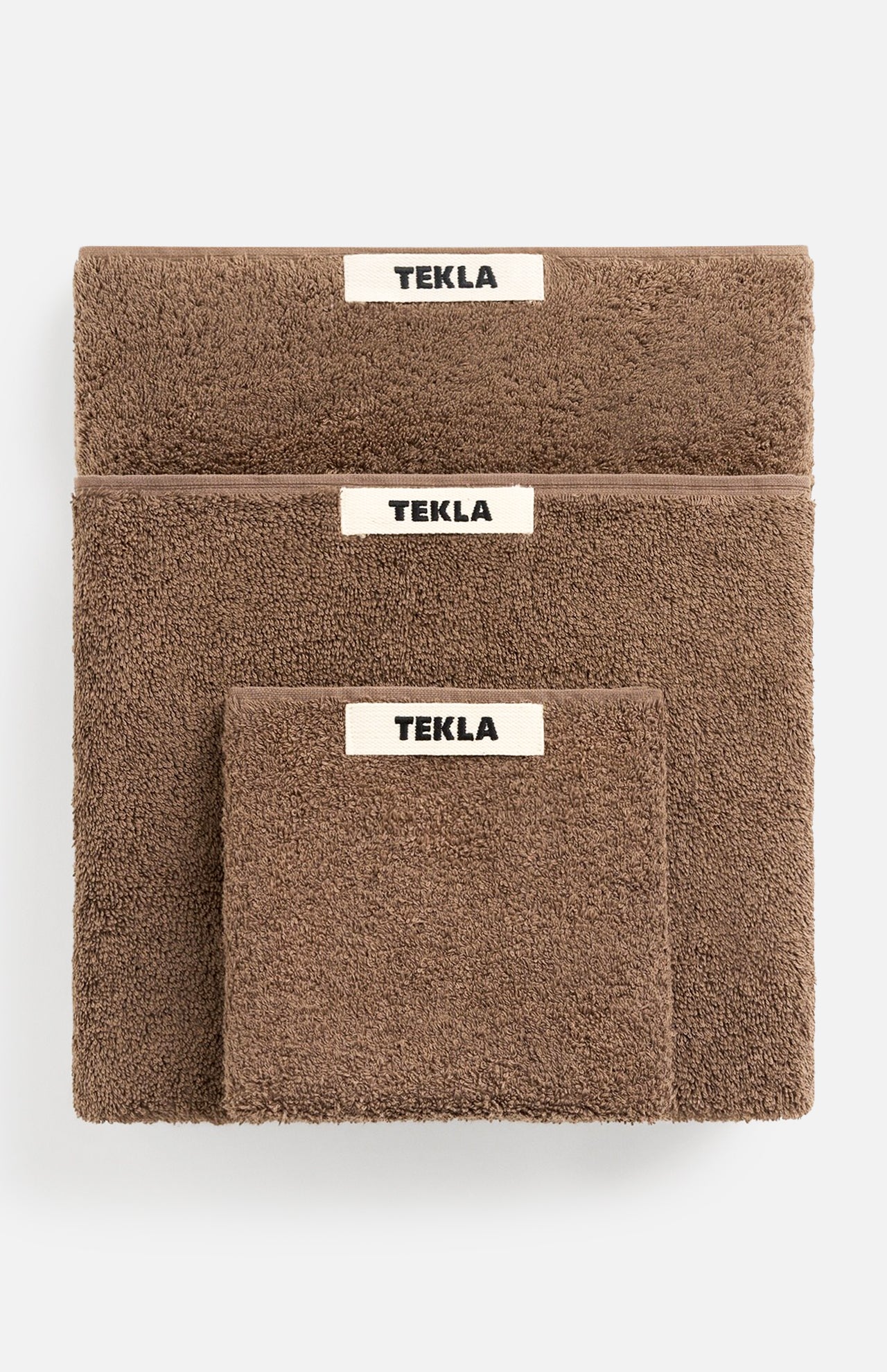 Three brown organic cotton TEKLA Terry Towel Washcloths of varying sizes stacked on top of each other, each featuring a white label with black text.