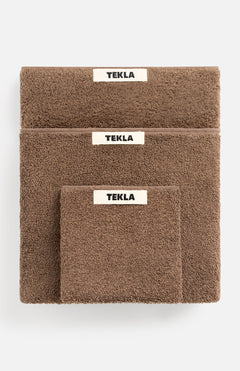 Three brown organic cotton TEKLA Terry Towel Washcloths of varying sizes stacked on top of each other, each featuring a white label with black text.