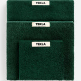A stack of dark green organic cotton Terry Towel Washcloths in varying sizes, each featuring a white TEKLA brand label.