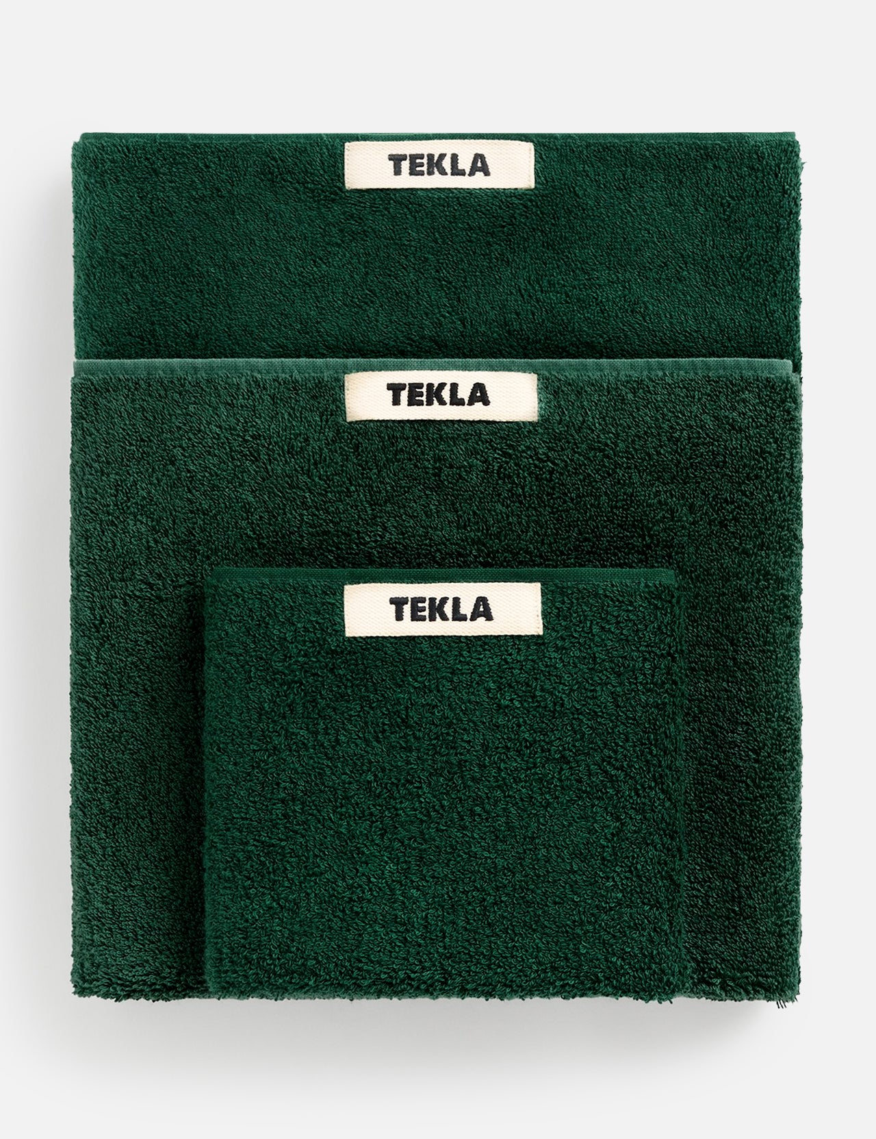 A stack of dark green organic cotton Terry Towel Washcloths in varying sizes, each featuring a white TEKLA brand label.