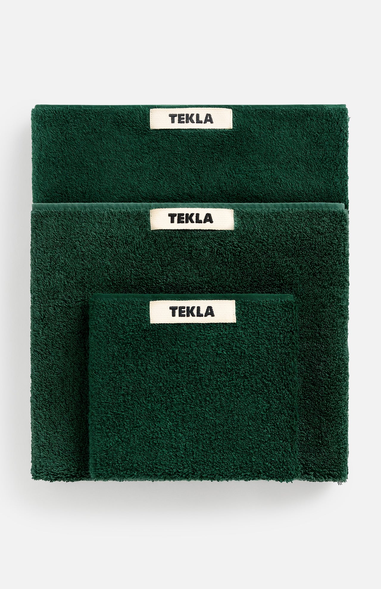 A stack of dark green organic cotton Terry Towel Washcloths in varying sizes, each featuring a white TEKLA brand label.
