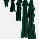 Five dark green organic cotton TEKLA Terry Towel Washcloths of varying sizes, including a soft bathroom towel and washcloth, hanging from hooks against a white wall.