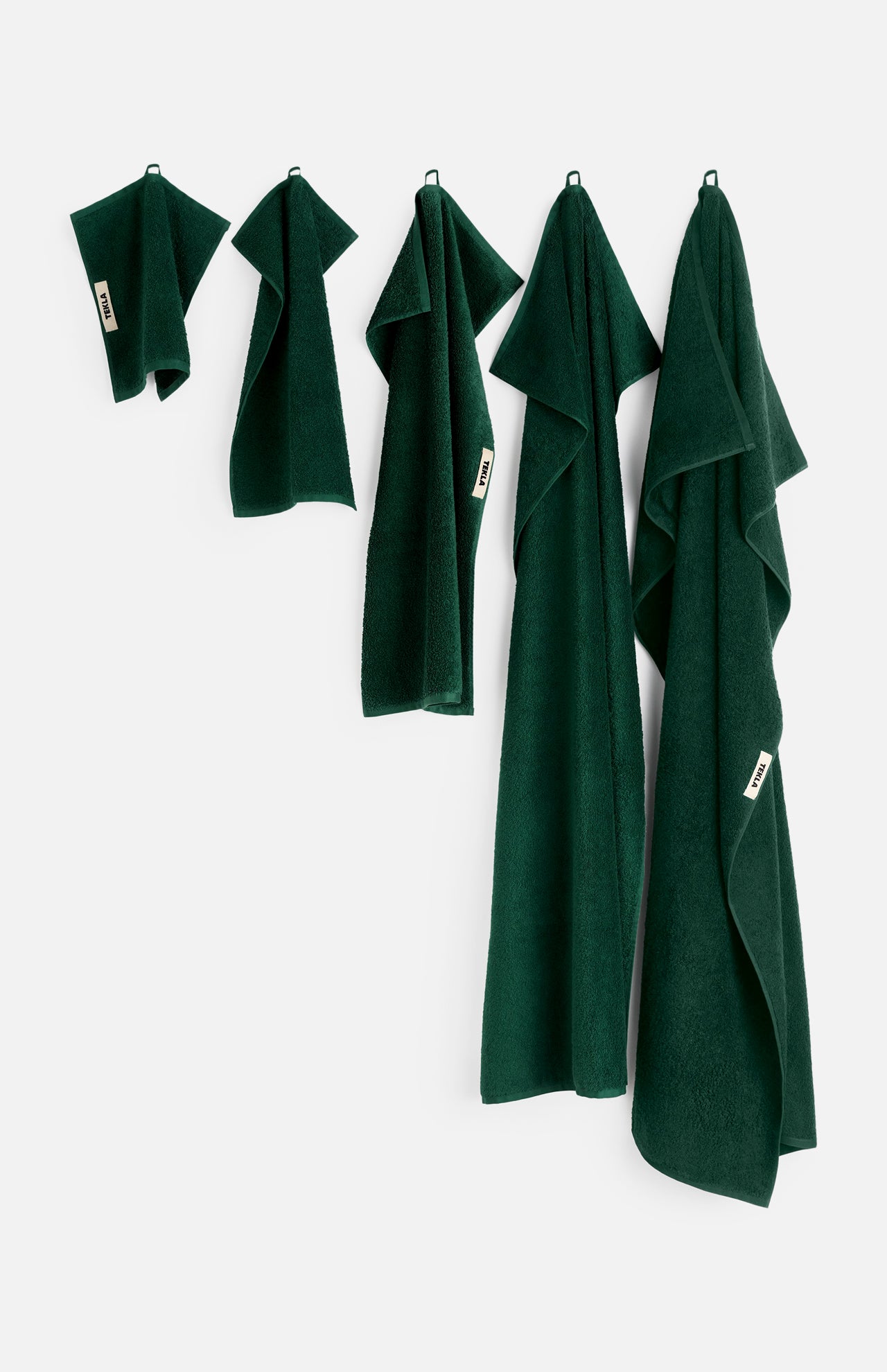 Five dark green organic cotton TEKLA Terry Towel Washcloths of varying sizes, including a soft bathroom towel and washcloth, hanging from hooks against a white wall.
