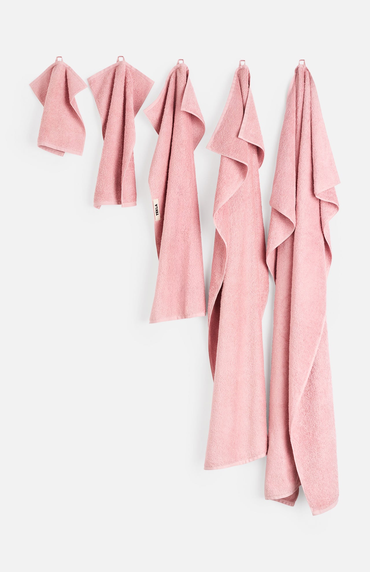 Five pink organic cotton Terry Towel Washcloths of varying sizes hanging in a row against a white wall.