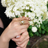 A person holds white hydrangeas, adorned with LOGAN HOLLOWELL’s French Pavé Diamond Tusk Ring alongside gold rings with pearls and a pearl bracelet. Their nails are manicured, and a soft blur of greenery fills the background.