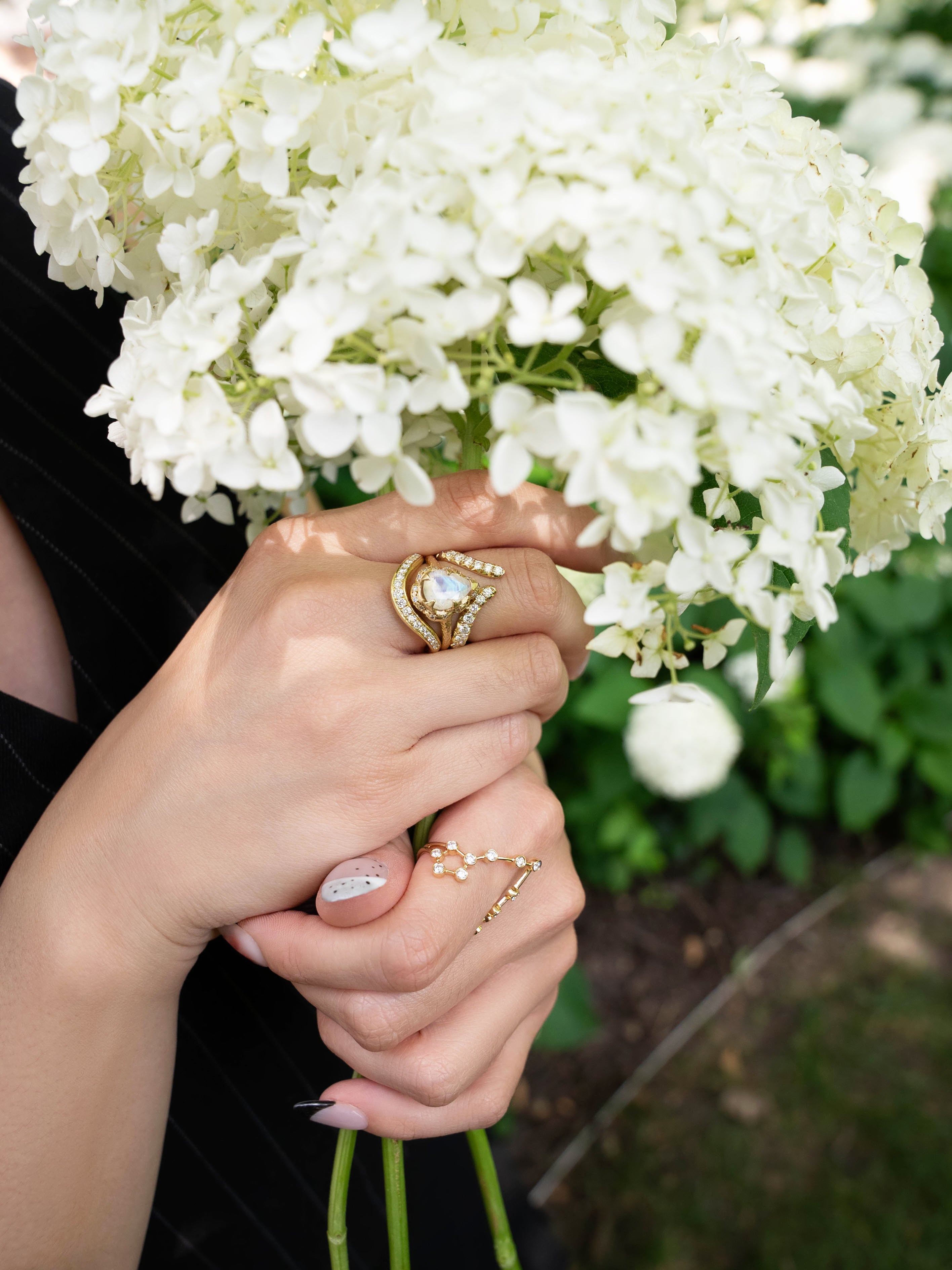 Hands holding a bouquet of white hydrangeas, adorned with gold rings featuring pearls and gems, including the LOGAN HOLLOWELL Pisces Diamond Constellation Ring. Nails are manicured, with one nail painted light pink and decorated with small white dots. Greenery is visible in the background.