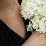 A person dressed in a black pinstripe outfit holds a bouquet of white flowers while wearing the LOGAN HOLLOWELL Gemini Diamond Constellation Necklace, featuring a yellow gold chain and diamond-accented constellation pendant.