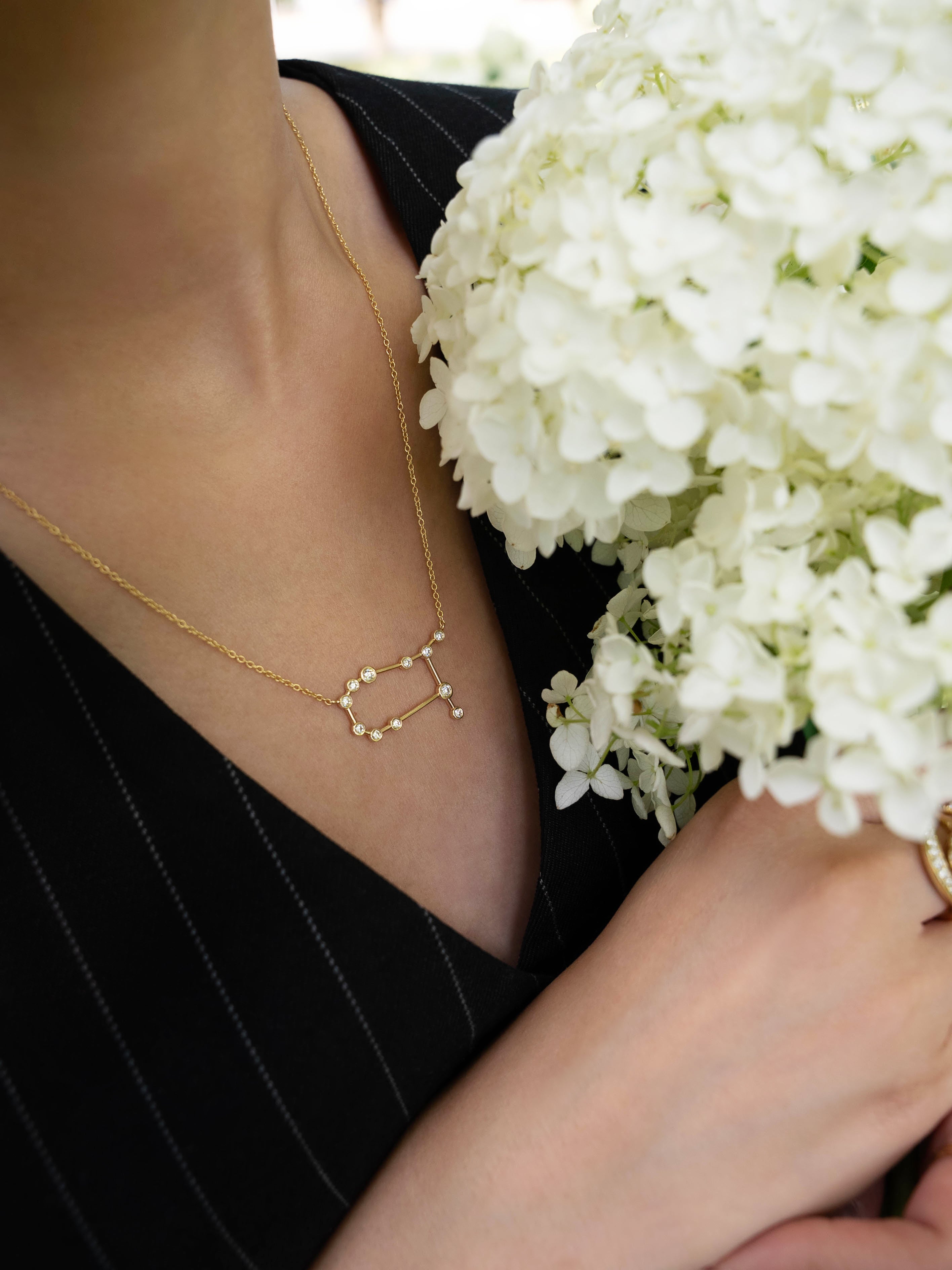 A person dressed in a black pinstripe outfit holds a bouquet of white flowers while wearing the LOGAN HOLLOWELL Gemini Diamond Constellation Necklace, featuring a yellow gold chain and diamond-accented constellation pendant.