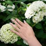 A hand with manicured nails adorned with the LOGAN HOLLOWELL Pisces Diamond Constellation Ring gently touches a cluster of white hydrangea flowers nestled among green leaves.