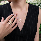 A person wearing a black pinstripe dress showcases the LOGAN HOLLOWELL Gemini Diamond Constellation Necklace, crafted in yellow gold with diamond accents, along with a textured gold ring. Their nails are manicured in a neutral design, and greenery is softly blurred in the background.
