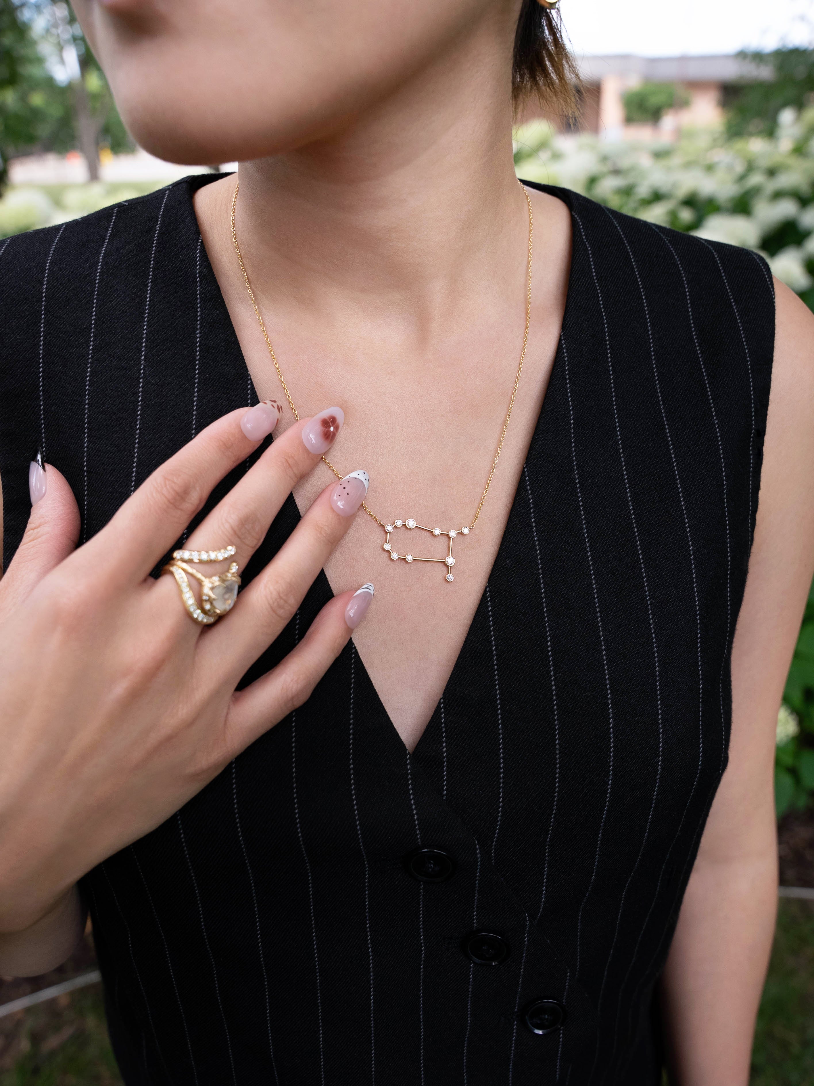 A person wearing a black pinstripe dress showcases the LOGAN HOLLOWELL Gemini Diamond Constellation Necklace, crafted in yellow gold with diamond accents, along with a textured gold ring. Their nails are manicured in a neutral design, and greenery is softly blurred in the background.