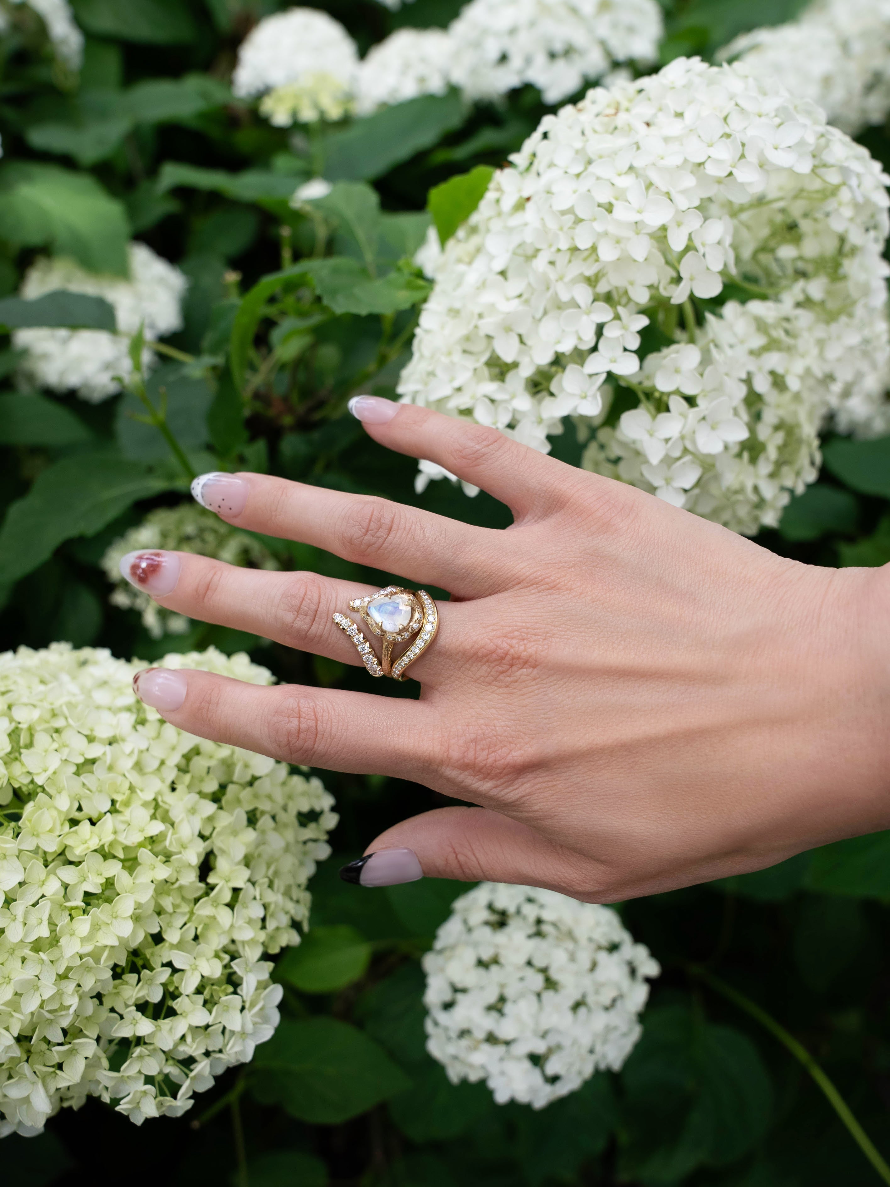 A hand with manicured nails and the LOGAN HOLLOWELL French Pavé Diamond Tusk Ring in gleaming 14k yellow gold rests gently on white hydrangea flowers in a garden.