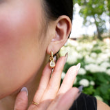 A person with long nails touches their ear, showcasing the LOGAN HOLLOWELL Enigma Diamond Oracle Set Water Drop Earrings in elegant 18k yellow gold. The background features softly blurred white flowers and green foliage.