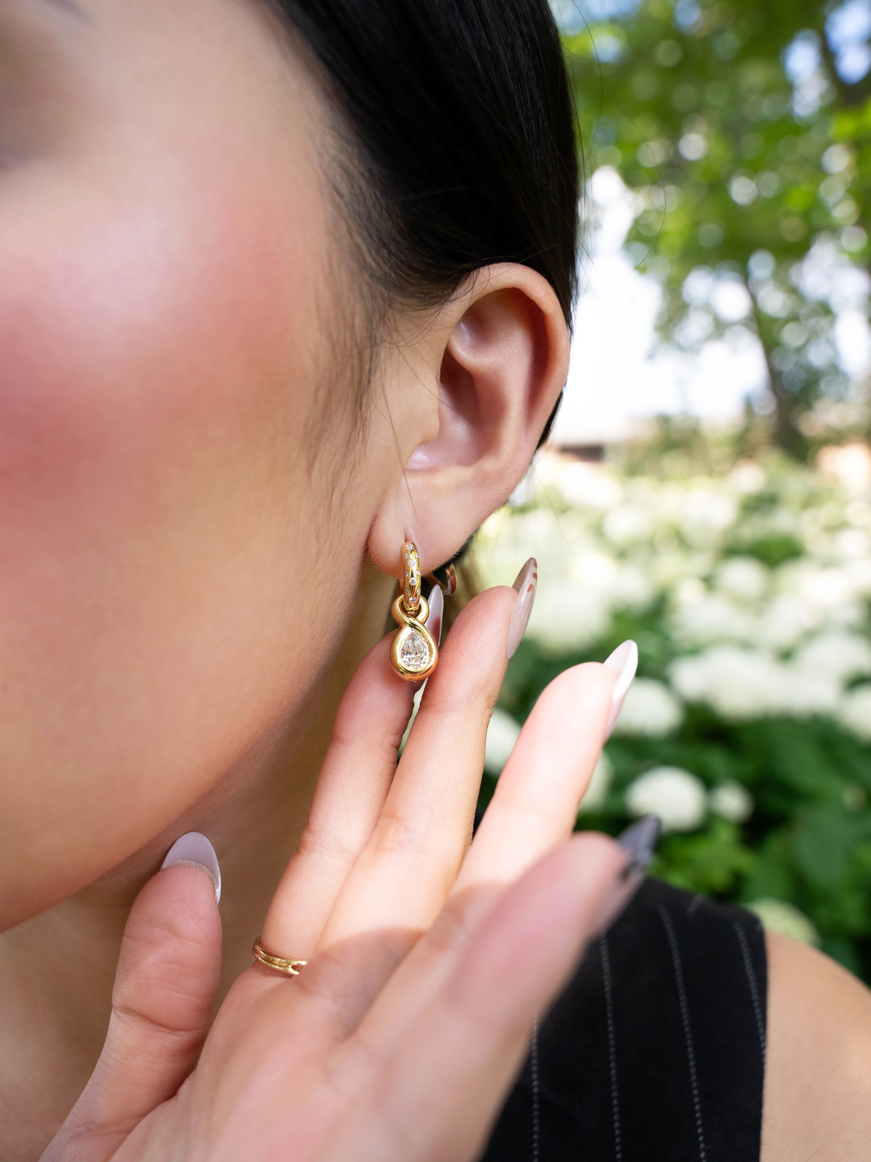 A person with long nails touches their ear, showcasing the LOGAN HOLLOWELL Enigma Diamond Oracle Set Water Drop Earrings in elegant 18k yellow gold. The background features softly blurred white flowers and green foliage.