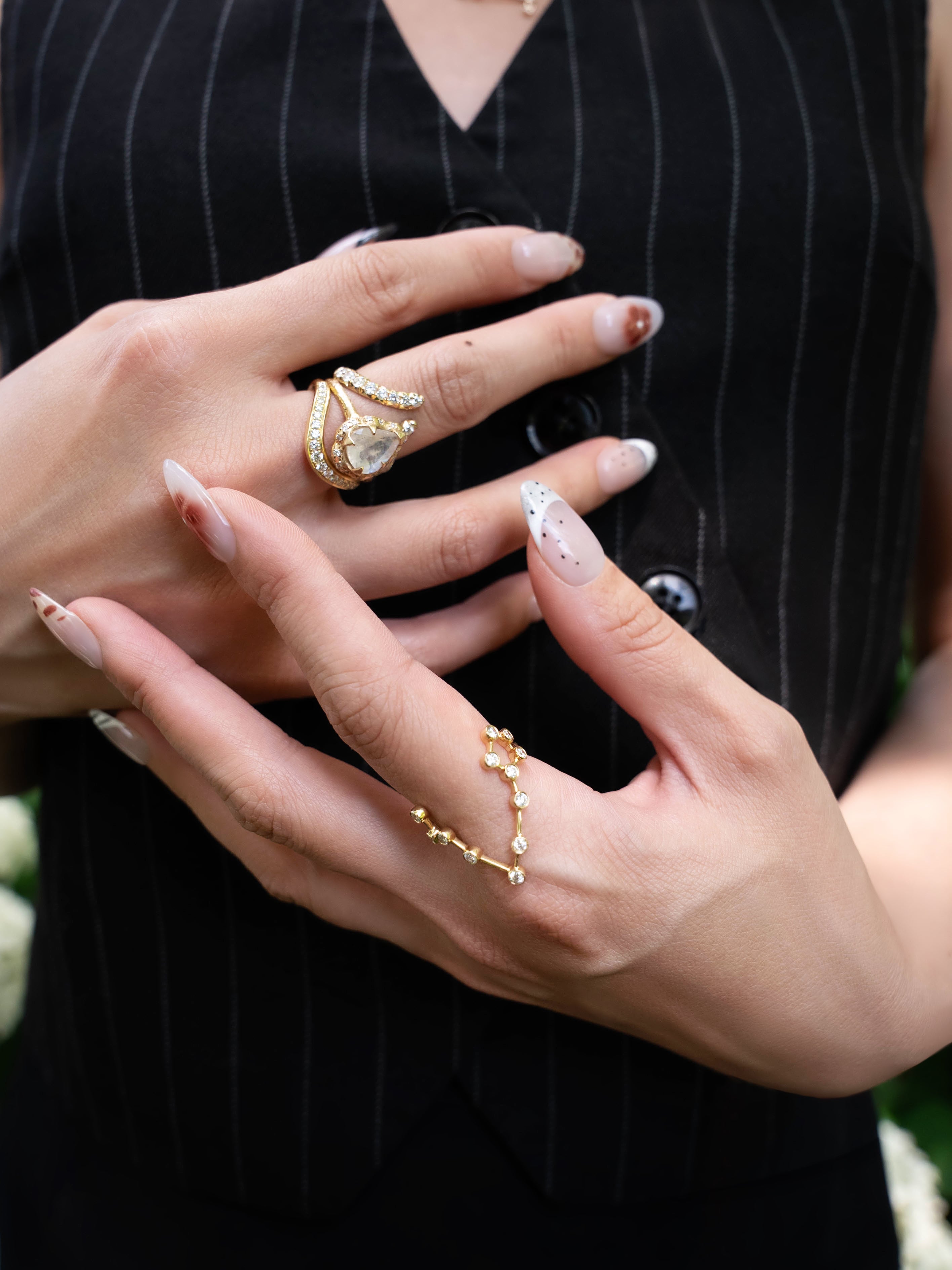 Close-up of a person’s hands with long, decorated nails adorned with multiple gold rings—including the LOGAN HOLLOWELL Pisces Diamond Constellation Ring—set against their black pinstriped vest. The image highlights the jewelry and manicure, drawing attention to the elegant zodiac-inspired design of the diamond constellation ring.