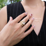 A person wearing a pinstripe vest, a gold constellation necklace, and several pieces of designer jewelry—including LOGAN HOLLOWELL’s Enigma Channel Band Ring in 18k yellow gold, alongside other gem-studded rings—rests their manicured hand on their chest. The delicate Enigma Channel Band Ring catches the light amid the collection. Blurred greenery is visible in the background.