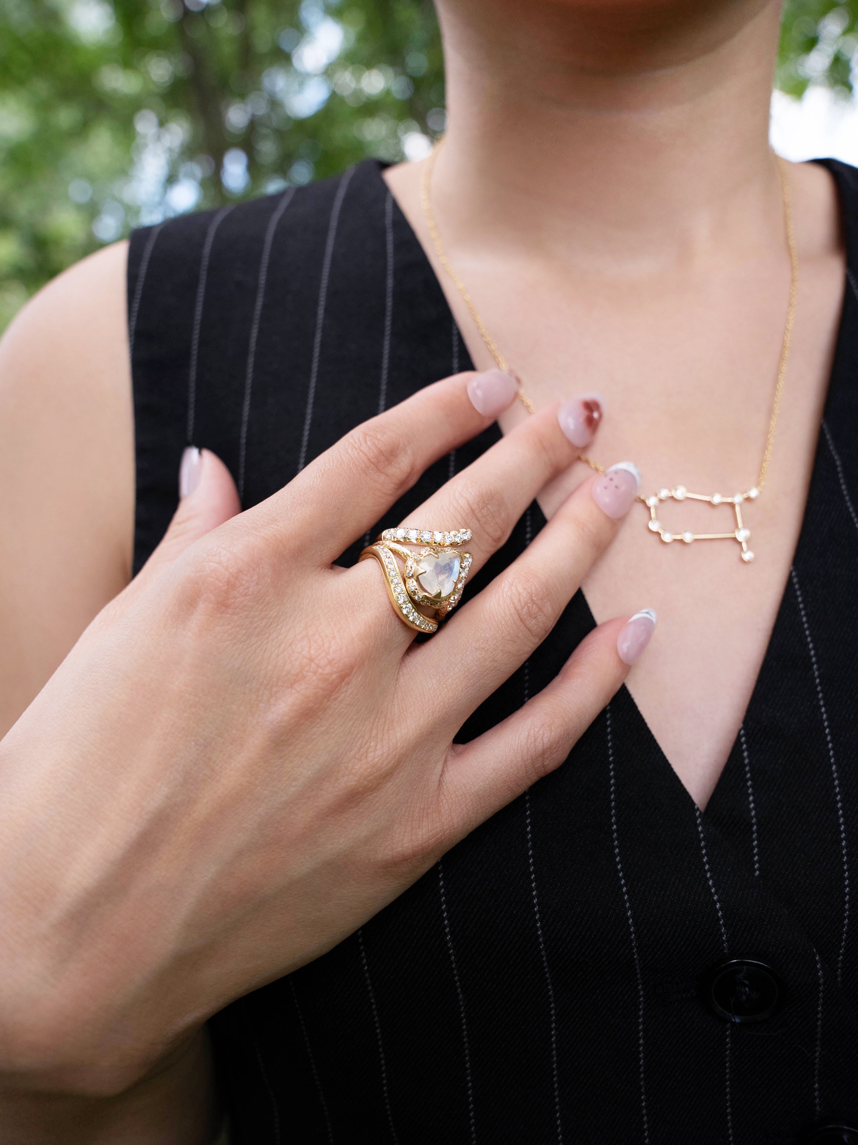 A person wearing a pinstripe vest, a gold constellation necklace, and several pieces of designer jewelry—including LOGAN HOLLOWELL’s Enigma Channel Band Ring in 18k yellow gold, alongside other gem-studded rings—rests their manicured hand on their chest. The delicate Enigma Channel Band Ring catches the light amid the collection. Blurred greenery is visible in the background.