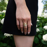 A close-up of a person’s hand wearing the LOGAN HOLLOWELL French Pavé Diamond Tusk Ring, resting against their thigh. They are dressed in a black skirt and standing in front of green bushes with white flowers.