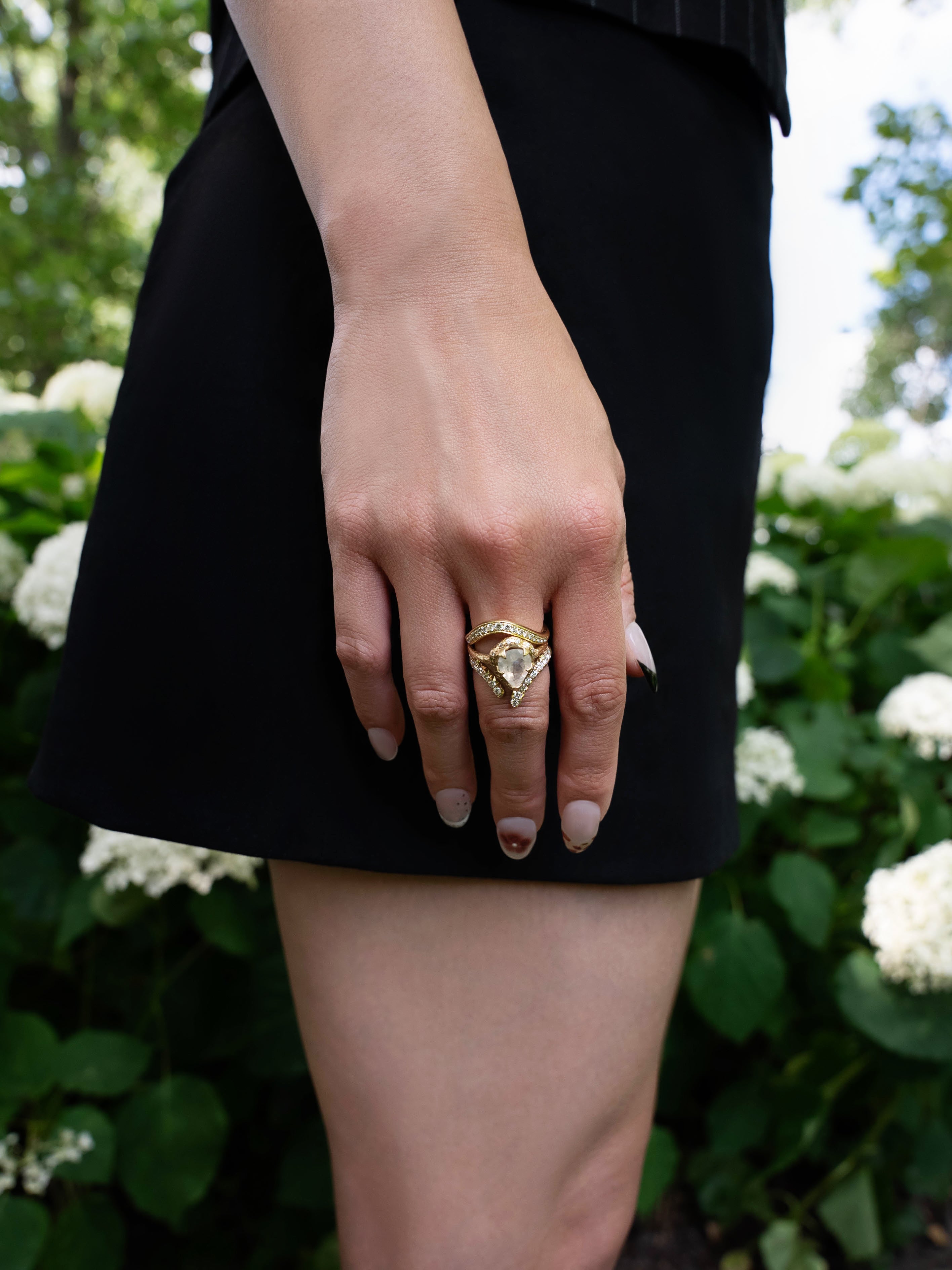 A close-up of a person’s hand wearing the LOGAN HOLLOWELL French Pavé Diamond Tusk Ring, resting against their thigh. They are dressed in a black skirt and standing in front of green bushes with white flowers.