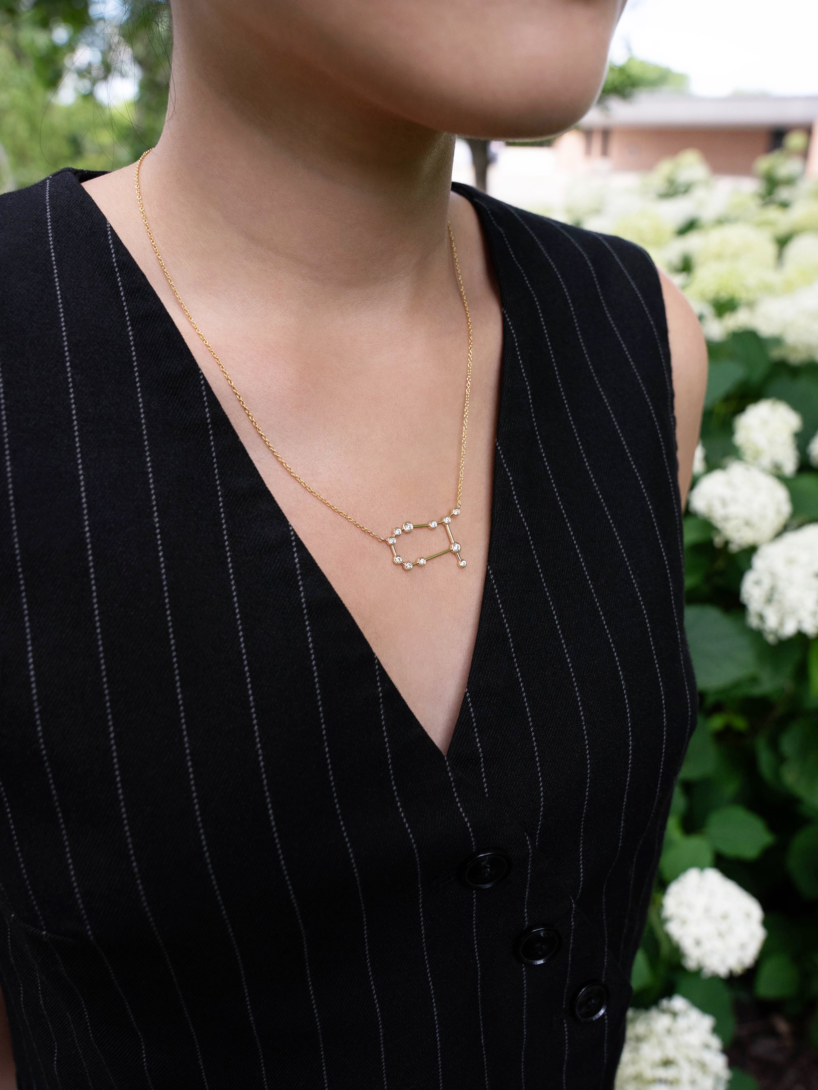 A person wearing a black pinstripe vest and the LOGAN HOLLOWELL Gemini Diamond Constellation Necklace—a yellow gold piece featuring a delicate constellation pendant with diamond accents—stands in front of white hydrangea flowers, elegantly showcasing this signature jewelry.