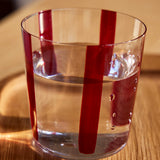 The Nova Stripe Tumbler from THE CONRAN SHOP, featuring vertical red stripes, is filled with water and placed on a wooden surface in natural sunlight.