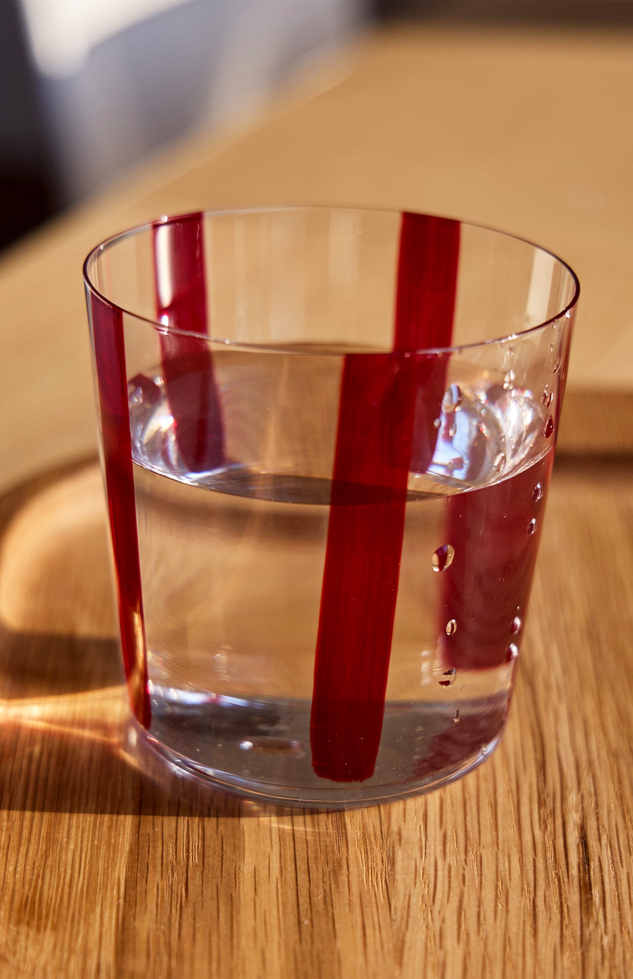 The Nova Stripe Tumbler from THE CONRAN SHOP, featuring vertical red stripes, is filled with water and placed on a wooden surface in natural sunlight.