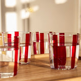 The Nova Stripe Tumbler Set of 4 by THE CONRAN SHOP features crystal glasses with vertical red stripes, casting striking shadows and light patterns on a wooden surface in warm sunlight.