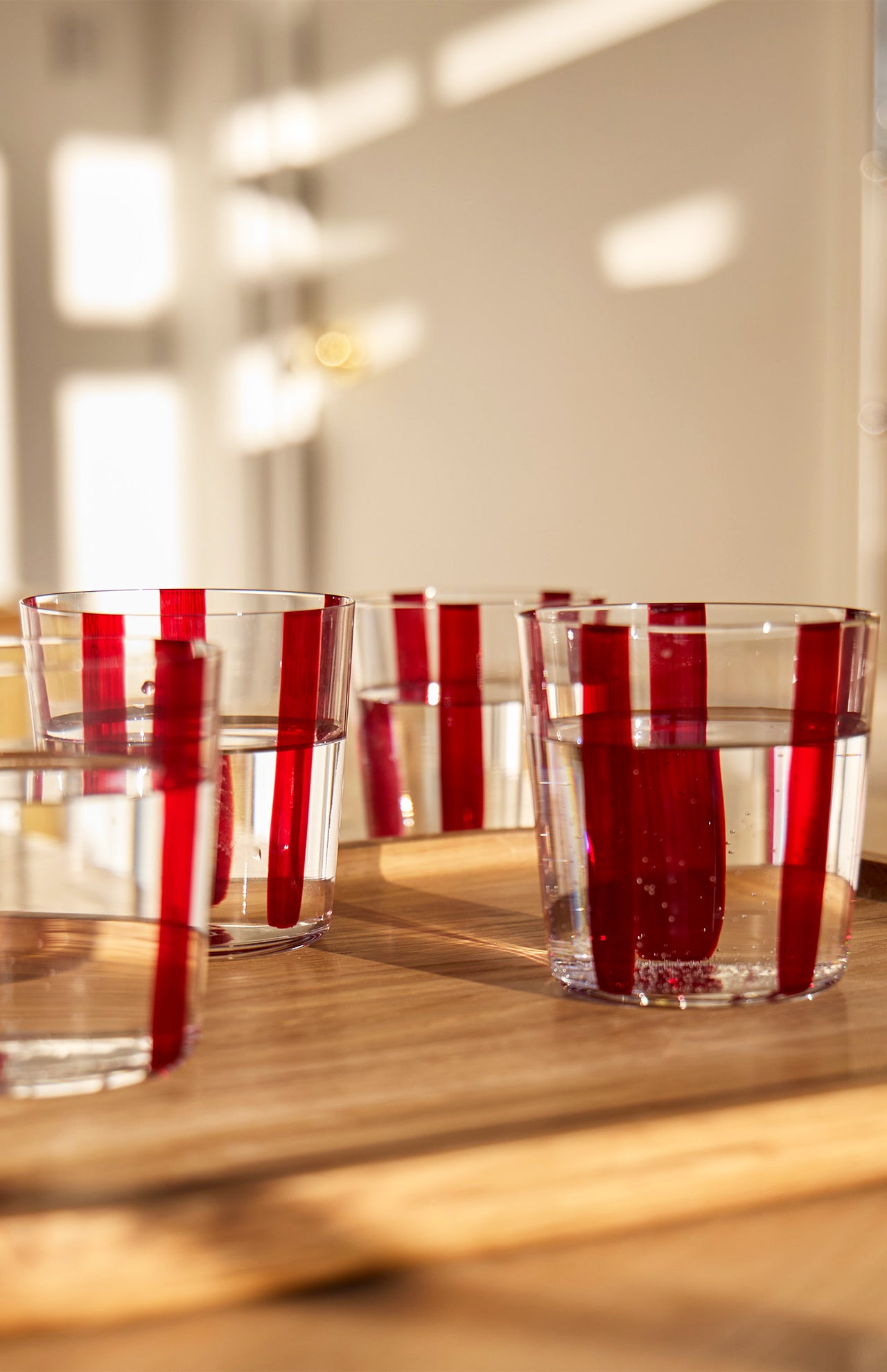 The Nova Stripe Tumbler Set of 4 by THE CONRAN SHOP features crystal glasses with vertical red stripes, casting striking shadows and light patterns on a wooden surface in warm sunlight.