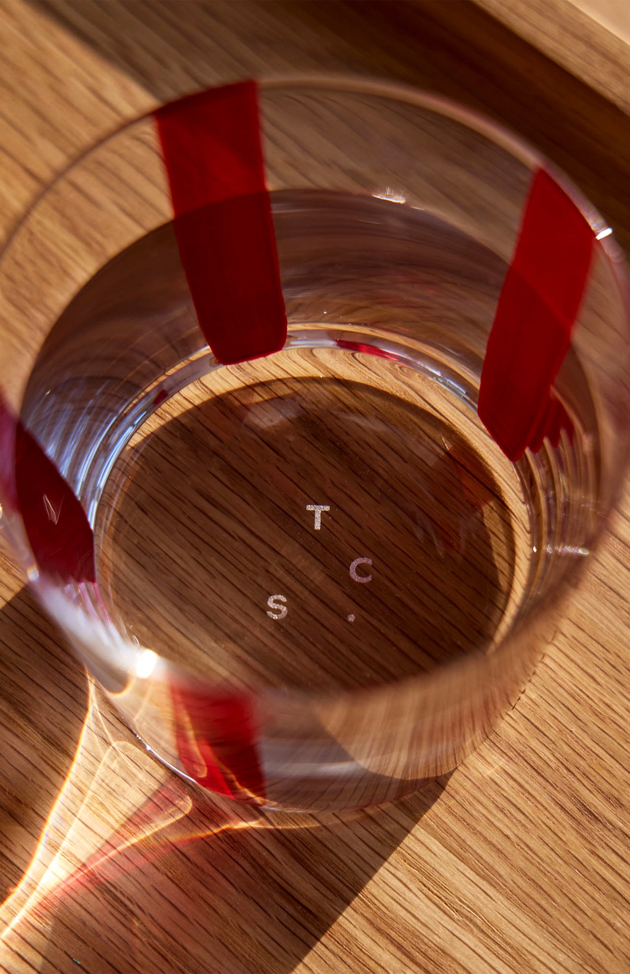 A Nova Stripe Tumbler from THE CONRAN SHOP, with red vertical stripes and water inside, sits on a wooden surface as sunlight reflects. The letters T, C, S, and C appear at the base of this striped glass from the set of 4.