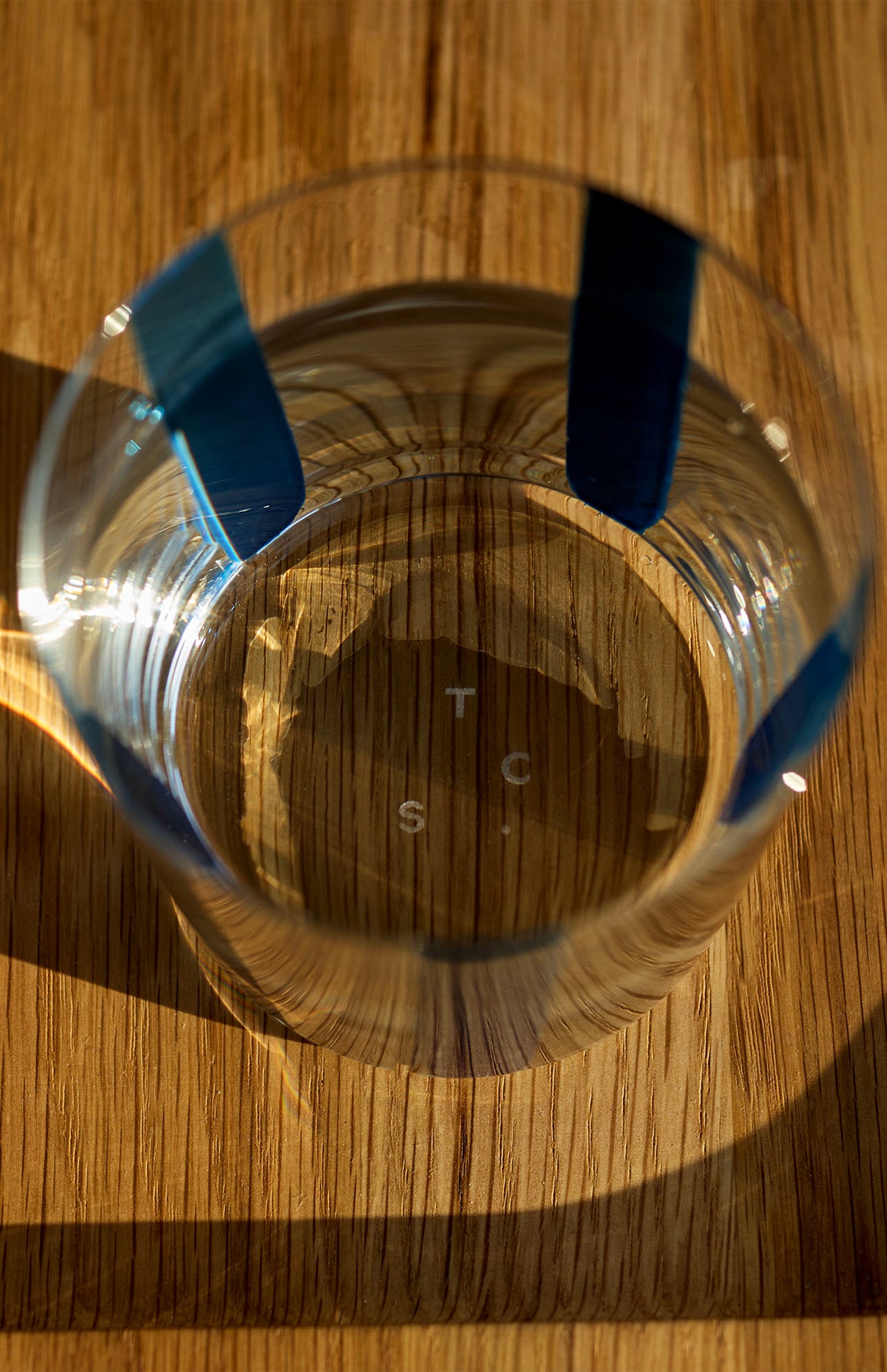 A hand-painted Nova Stripe Tumbler from THE CONRAN SHOP, featuring blue vertical stripes, rests on a wooden surface and casts a shadow. The crystal glass base is marked with the letters “T,” “S,” and “C.”.