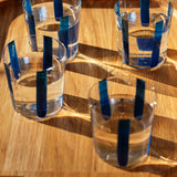 The Nova Stripe Tumbler Set of 4 by THE CONRAN SHOP, featuring crystal glasses with vertical blue stripes, sits on a wooden tray as sunlight creates inviting shadows and reflections.