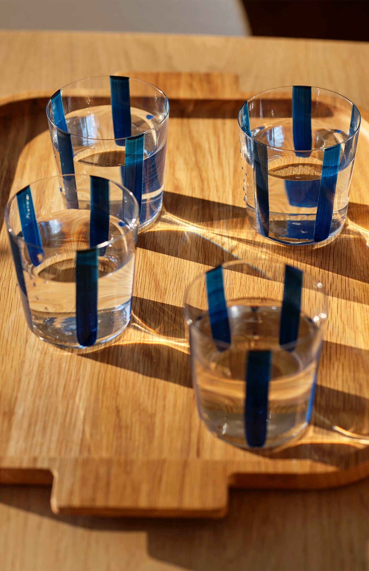 The Nova Stripe Tumbler Set of 4 by THE CONRAN SHOP, featuring crystal glasses with vertical blue stripes, sits on a wooden tray as sunlight creates inviting shadows and reflections.