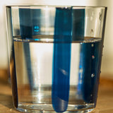 A clear glass of water on wood shows blue vertical reflections and shadows, echoing the Nova Stripe Tumbler Set of 4 by THE CONRAN SHOP—stripe patterns are visible through the liquid and on the glass.