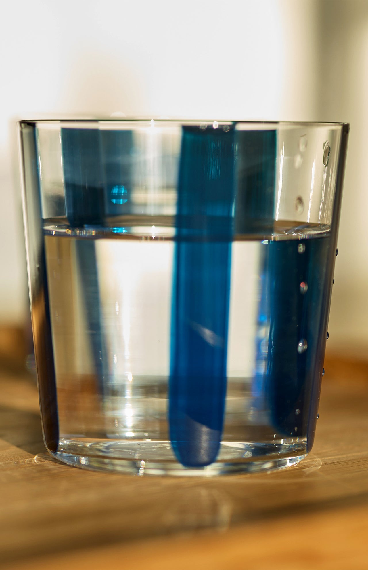 A clear glass of water on wood shows blue vertical reflections and shadows, echoing the Nova Stripe Tumbler Set of 4 by THE CONRAN SHOP—stripe patterns are visible through the liquid and on the glass.