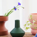 The Sphere Bottle Vase by THE CONRAN SHOP in forest green sits on a wooden surface between a brown pot with blue flowers and a pink pot with white flowers, set against a softly blurred light background.