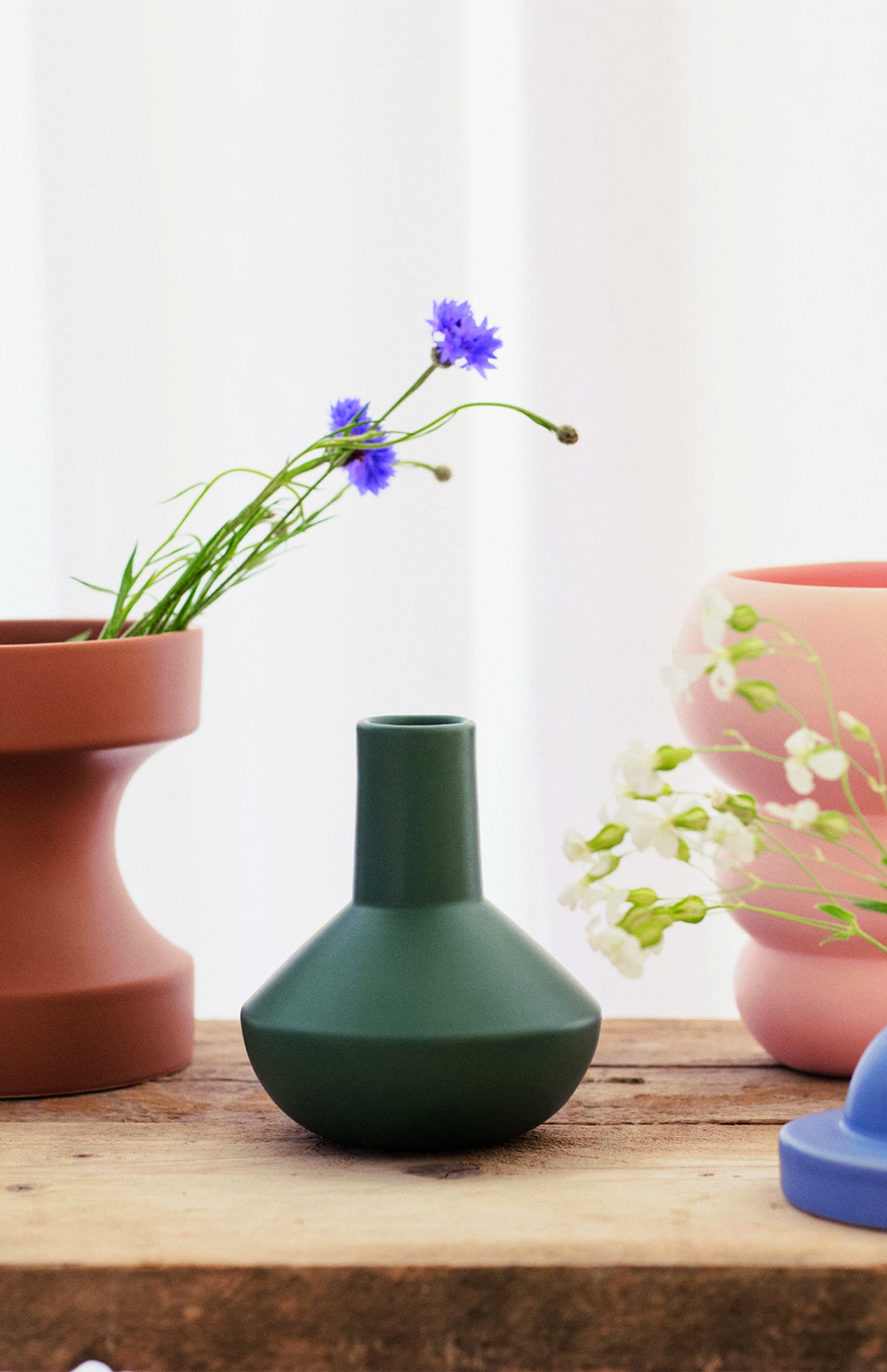 The Sphere Bottle Vase by THE CONRAN SHOP in forest green sits on a wooden surface between a brown pot with blue flowers and a pink pot with white flowers, set against a softly blurred light background.