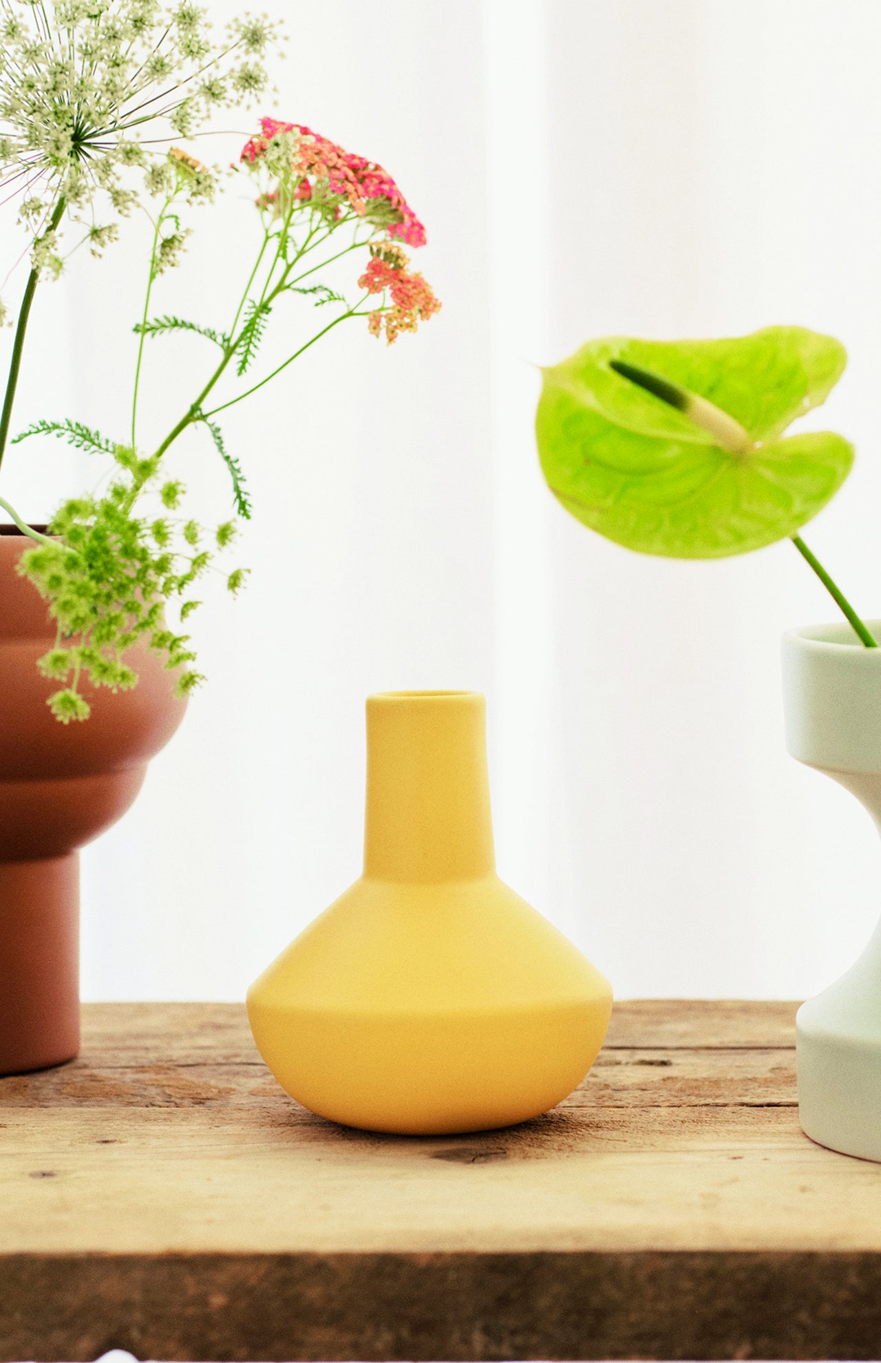 A yellow Sphere Bottle Vase by THE CONRAN SHOP is displayed on a wooden surface between sculptural vases with green and pink flowers, set against a soft white curtain in the background.