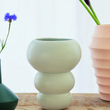 The Stacked Spheres Vase Mint by THE CONRAN SHOP rests on a wooden surface, joined by other distinctively shaped earthenware vases with flowers, all set against a softly lit white background.