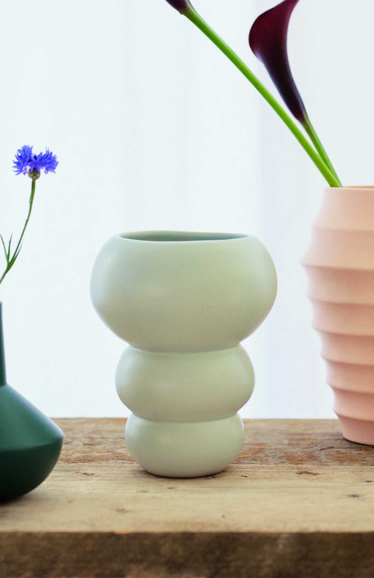 The Stacked Spheres Vase Mint by THE CONRAN SHOP rests on a wooden surface, joined by other distinctively shaped earthenware vases with flowers, all set against a softly lit white background.