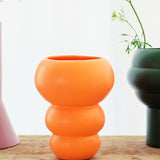 The Stacked Spheres Vase Orange by THE CONRAN SHOP sits on wood, with a glimpse of a purple vase to the left and a green earthenware vase with delicate greenery to the right, all set against a light background.