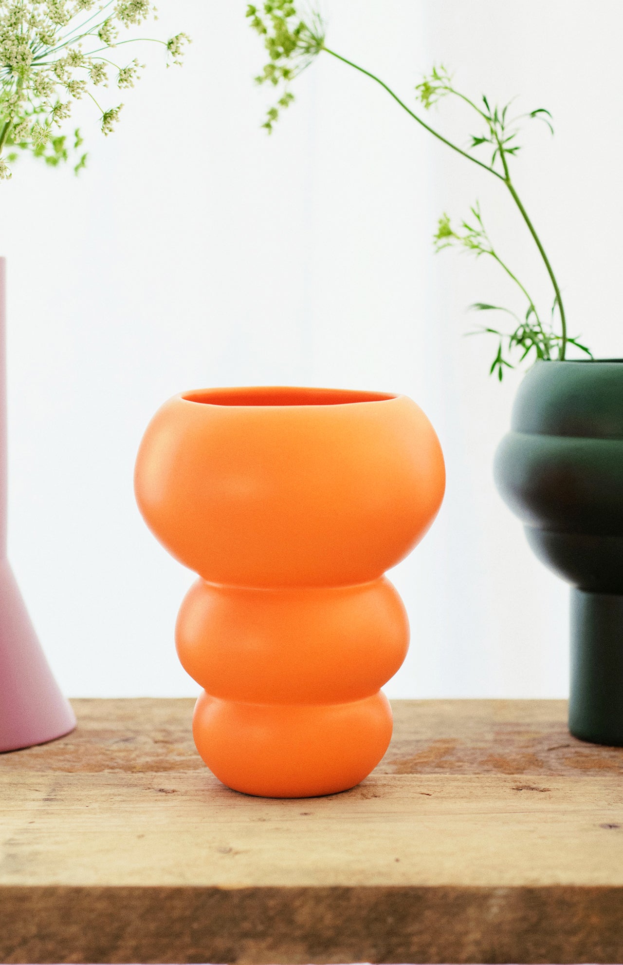 The Stacked Spheres Vase Orange by THE CONRAN SHOP sits on wood, with a glimpse of a purple vase to the left and a green earthenware vase with delicate greenery to the right, all set against a light background.