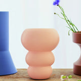 The Stacked Spheres Vase Plaster by THE CONRAN SHOP, a matte pale pink earthenware piece with a rounded stacked design, sits on wood between a tall blue vase and a brown flower planter, softly lit by a white curtain in the background.