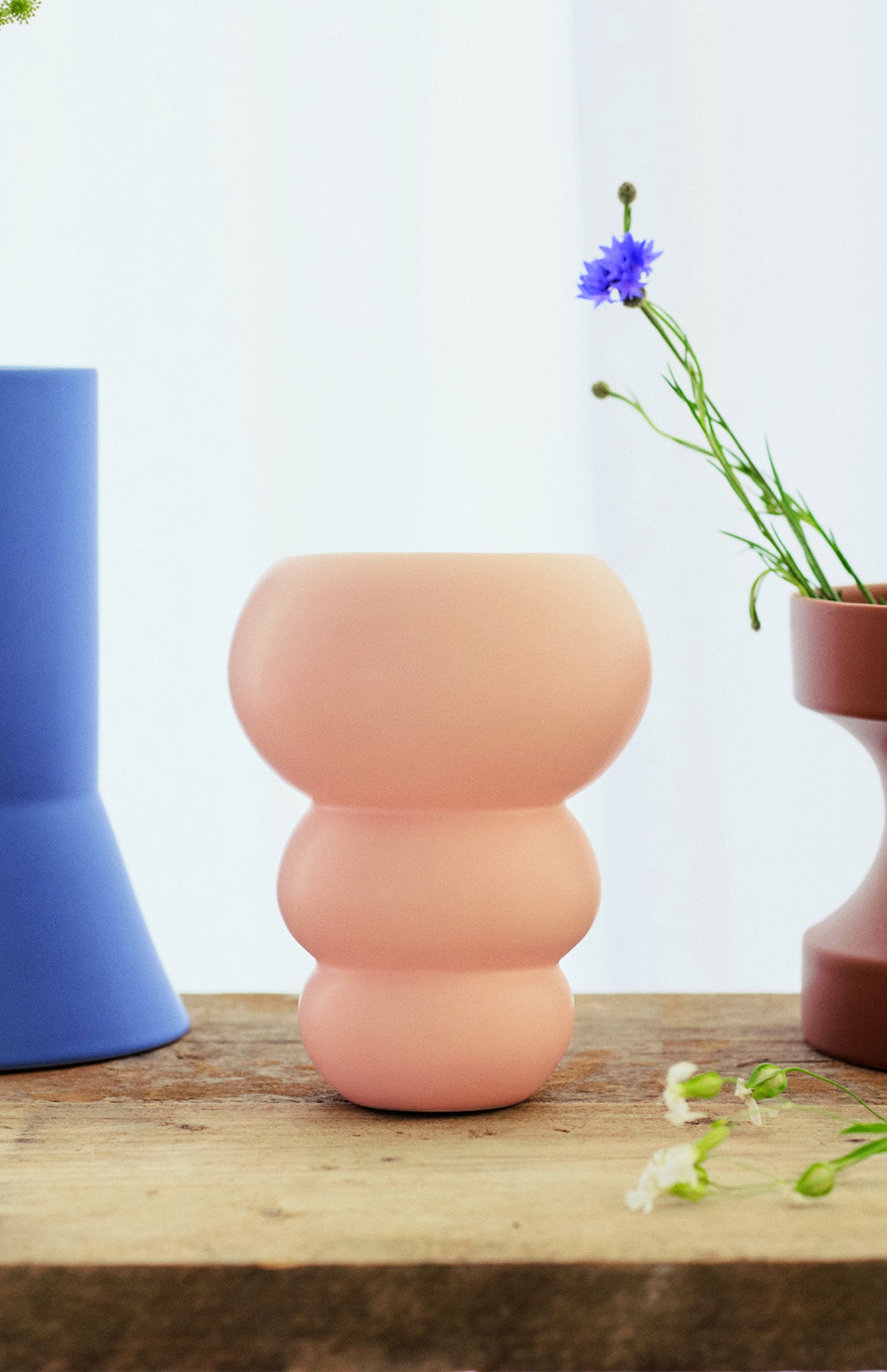 The Stacked Spheres Vase Plaster by THE CONRAN SHOP, a matte pale pink earthenware piece with a rounded stacked design, sits on wood between a tall blue vase and a brown flower planter, softly lit by a white curtain in the background.