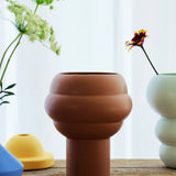 The Tree Vase Cocao by THE CONRAN SHOP, a modern brown earthenware piece with rounded, layered design, rests on a wooden surface amid colorful vases and fresh flowers, with a sheer curtain softly framing the scene.