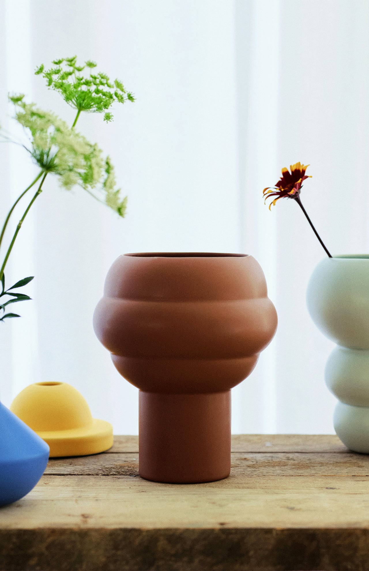 The Tree Vase Cocao by THE CONRAN SHOP, a modern brown earthenware piece with rounded, layered design, rests on a wooden surface amid colorful vases and fresh flowers, with a sheer curtain softly framing the scene.