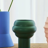 The Tree Vase Forest Green by THE CONRAN SHOP sits on a wooden surface, flanked by a light blue earthenware vase with green stems on the left and a peach ribbed vase on the right, against softly lit white curtains.