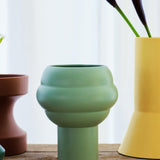 The Tree Vase Sage Green by THE CONRAN SHOP, with a rounded centre and matte finish, sits on wood among brown and yellow vases, dark purple flowers behind it, all set against a white curtain.