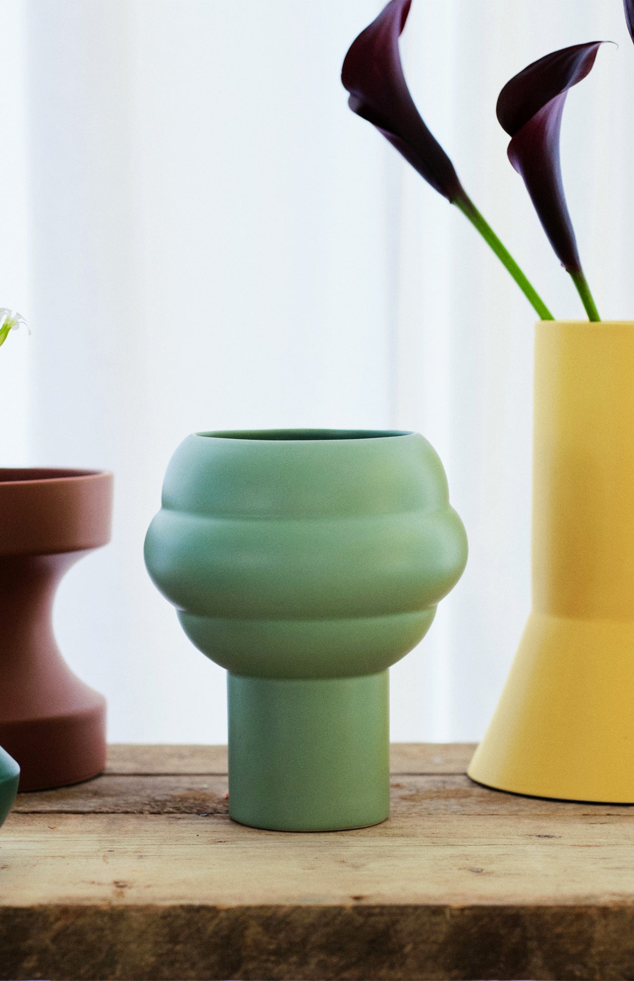 The Tree Vase Sage Green by THE CONRAN SHOP, with a rounded centre and matte finish, sits on wood among brown and yellow vases, dark purple flowers behind it, all set against a white curtain.