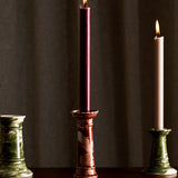 Three Marbled Wardour Candlesticks by THE CONRAN SHOP—one red with a lit burgundy candle, two green (one with an unlit white candle)—stand on a dark surface before a softly lit curtain. Each candlestick features hand-marbled glaze.